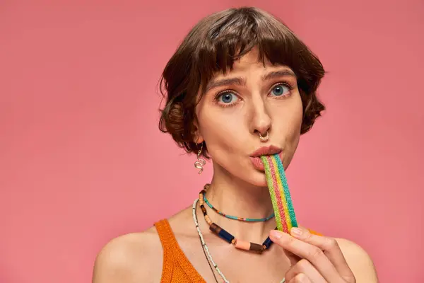 Portrait of playful woman in her 20s biting sweet and sour candy strip on pink background — Stock Photo