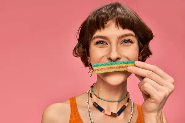 Portrait of woman in her 20s covering lips with sweet and sour candy strip on pink background — Stock Photo