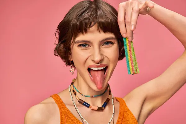 Happy woman in her 20s holding sweet and sour candy strip and sticking tongue on pink background — Stock Photo