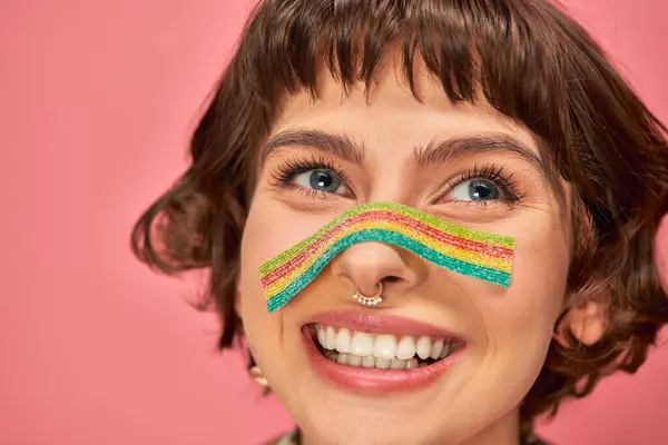 Happy woman in her 20s with sweet and sour candy strip aon her nose on pink background — Stock Photo
