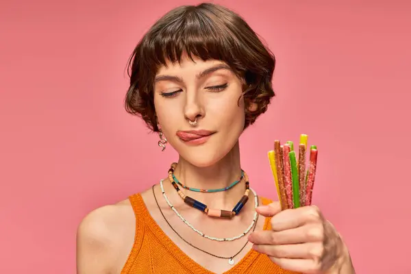 Juguetona jovencita de unos 20 años con lengua pegajosa y dulces y amargos en la mano sobre rosa - foto de stock