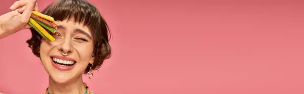 Joyeuse jeune femme dans la vingtaine souriante et tenant des bonbons aigre-doux à la main sur rose, bannière — Photo de stock