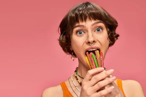 Смішна молода жінка в 20 років кладе купу солодких і кислих цукерок у відкритий рот на рожевий — стокове фото