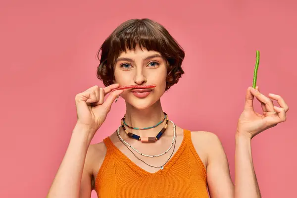 Chica juguetona en sus 20 años sonriendo y sosteniendo dulces y amargos sobre sus labios y bigote en rosa - foto de stock
