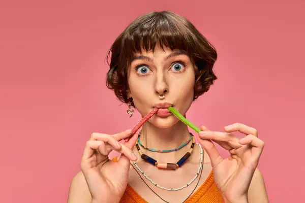 Menina perfurada engraçado em seus 20s comendo dois sabores diferentes de doces doces e azedos em rosa — Fotografia de Stock