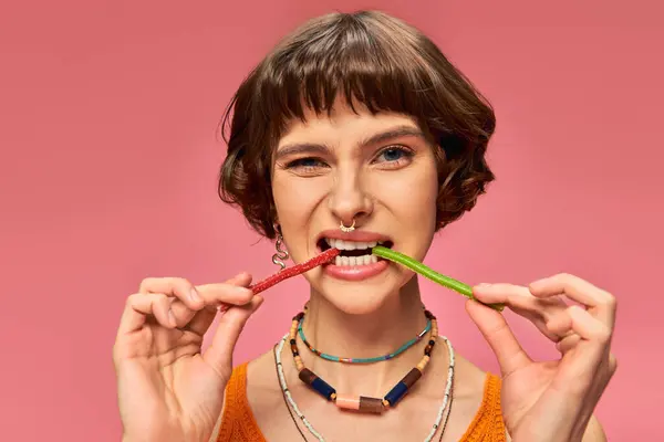 Menina perfurada engraçado em 20s mordendo dois sabores diferentes de doces doces e azedos no fundo rosa — Fotografia de Stock