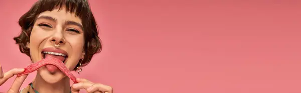 Glückliche junge Frau mit Nasenpiercing leckt süß-sauren Bonbonstreifen auf rosa Hintergrund, Banner — Stockfoto