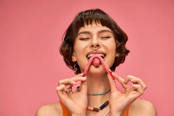 Довольная и молодая женщина с пирсингом носа облизывая сладкие и кислые конфеты полоску на розовом фоне — стоковое фото
