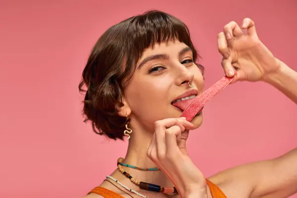 Веселая и молодая женщина с пирсингом носа лижет сладкие и кислые конфеты полоску на розовом фоне — стоковое фото