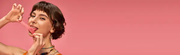 Glückliches Mädchen mit Nasenpiercing leckt süß-sauren Bonbonstreifen auf rosa Hintergrund, horizontal — Stockfoto