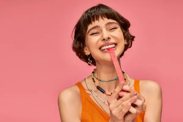 Aufgeregt und junge Frau mit Nasenpiercing beißenden süßen und sauren Bonbonstreifen auf rosa Hintergrund — Stockfoto