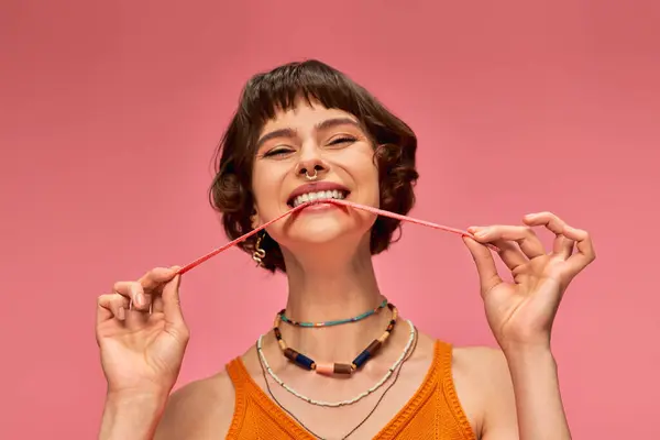 Восхитительная и молодая женщина с пирсингом носа кусает сладкие и кислые конфеты полоску на розовом фоне — стоковое фото