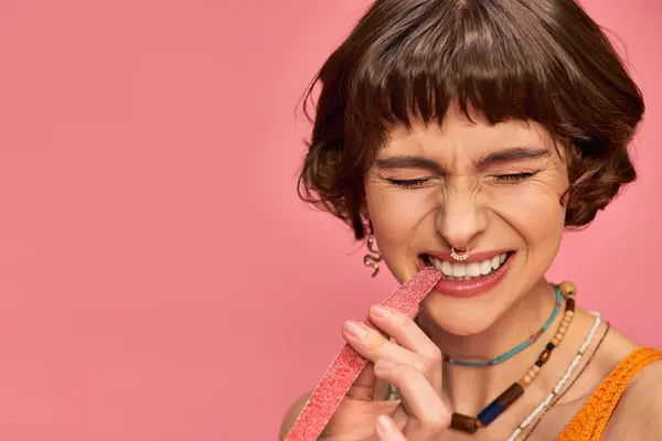 Экспрессивная женщина с белыми зубами и носом кусает сладкие и кислые конфеты полоски на розовый — стоковое фото
