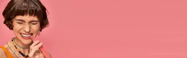 Fille expressive avec piercing et dents blanches mordant bande de bonbons aigre-douce sur rose, bannière — Photo de stock