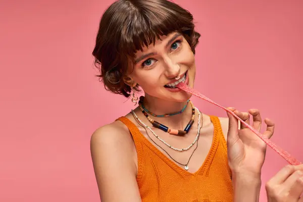 Милая девушка в 20 с пирсингом носа и белыми зубами кусая сладкие и кислые конфеты полоски, розовый набор — стоковое фото