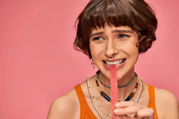 Menina emocional em 20s com piercing no nariz e dentes brancos mordendo doce e azedo tira de doces — Fotografia de Stock