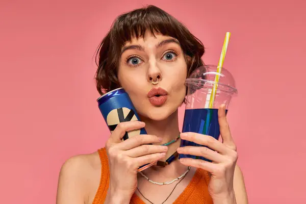 Amazed young woman with short brunette hair and piercing holding summer drinks on pink backdrop — Stock Photo
