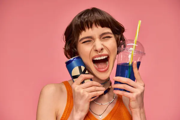Excited young woman with short brunette hair and piercing holding summer drinks on pink backdrop — Stock Photo
