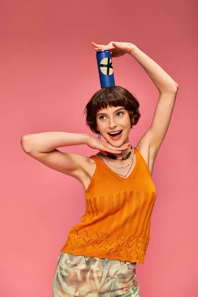 Feliz joven con pelo corto morena posando con lata de refresco en la cabeza en rosa, bebida de verano - foto de stock