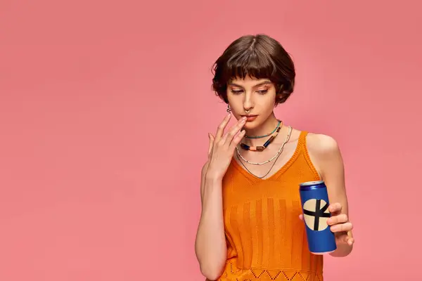 Doubtful young woman with short brunette hair looking at her soda can on pink, summer drink — Stock Photo