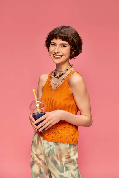 Весёлая молодая женщина с короткими волосами брюнетки позирует с освежающим летним коктейлем на розовом — стоковое фото