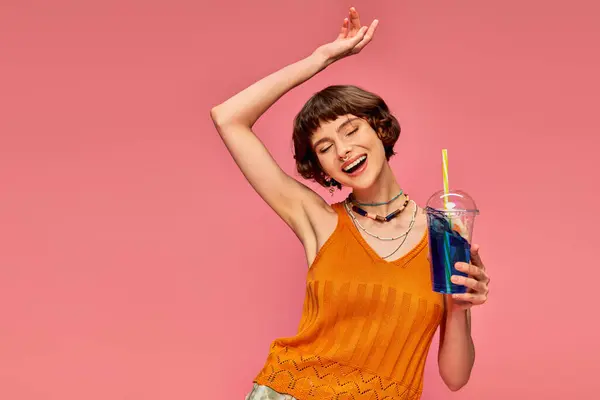 Excited young woman with short brunette hair holding refreshing summer drink over head on pink — Stock Photo