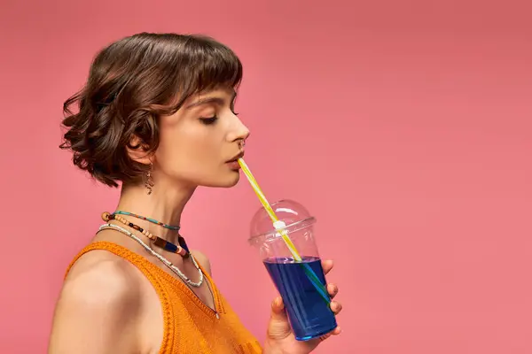 Young woman with short brunette hair drinking refreshing summer cocktail from straw on pink — Stock Photo