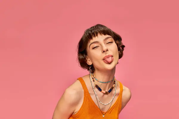 Cheeky carefree girl in 20s with short brunette hair sticking tongue out on pink background — Stock Photo