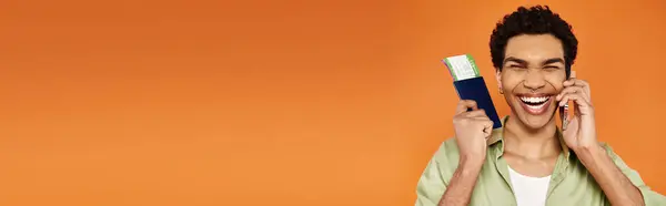 Joyful african american man in cozy attire talking by phone and holding passport with ticket, banner — Stock Photo