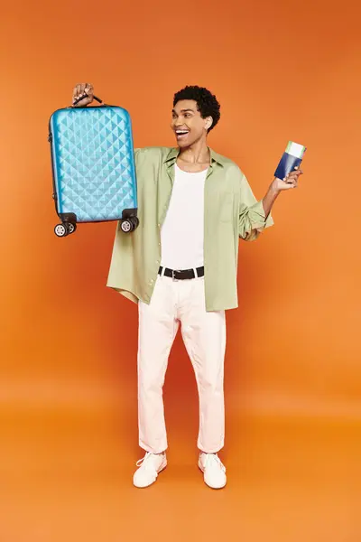 Atractivo hombre afroamericano alegre con pasaporte y billete y mirando la maleta azul - foto de stock