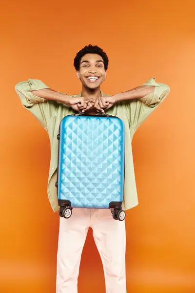 Alegre guapo africano americano hombre en casual traje sosteniendo azul maleta y sonriendo a la cámara - foto de stock