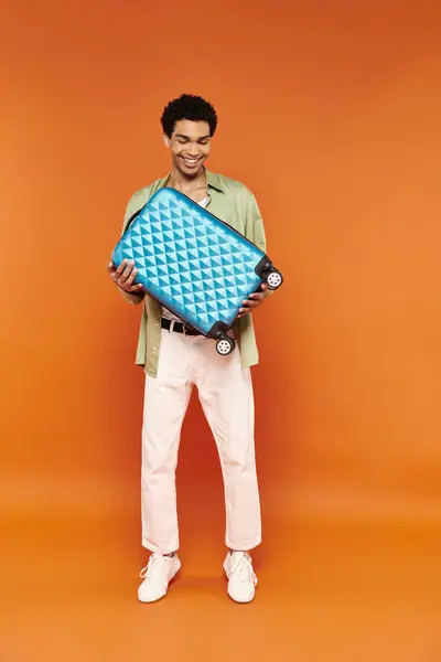Alegre africano americano hombre en traje casual mirando azul maleta en manos en naranja telón de fondo - foto de stock