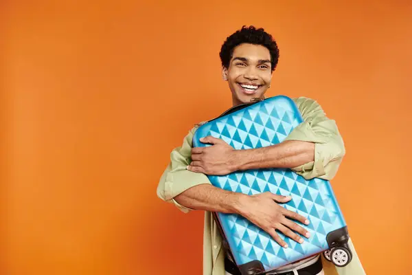Guapo y alegre hombre afroamericano con elegante atuendo abrazando su maleta azul sobre fondo naranja - foto de stock