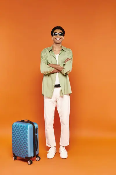 Joyeux homme afro-américain en tenue décontractée avec des lunettes de soleil élégantes posant à côté de la valise bleue — Photo de stock