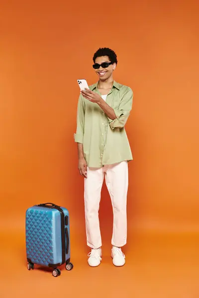 Allegro uomo afro americano con eleganti occhiali da sole in posa vicino a valigia blu e guardando il telefono — Foto stock