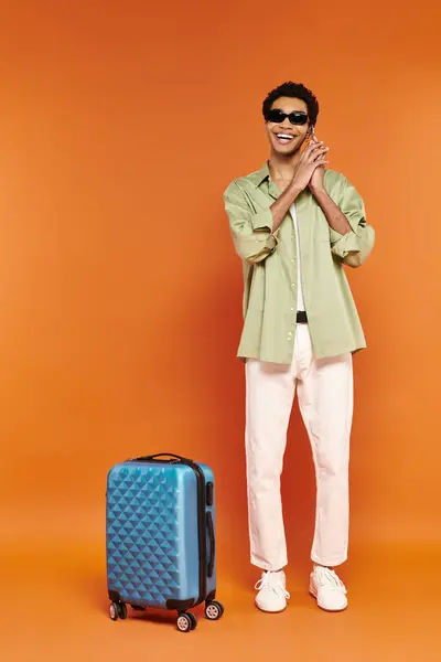 Atractivo hombre afroamericano feliz con gafas de sol de moda hablando por teléfono cerca de la maleta azul - foto de stock