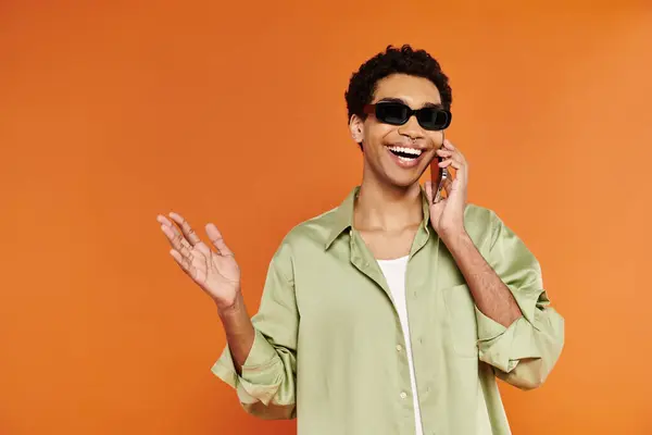 Bell'uomo afro-americano allegro con eleganti occhiali da sole che parlano per telefono su sfondo arancione — Foto stock