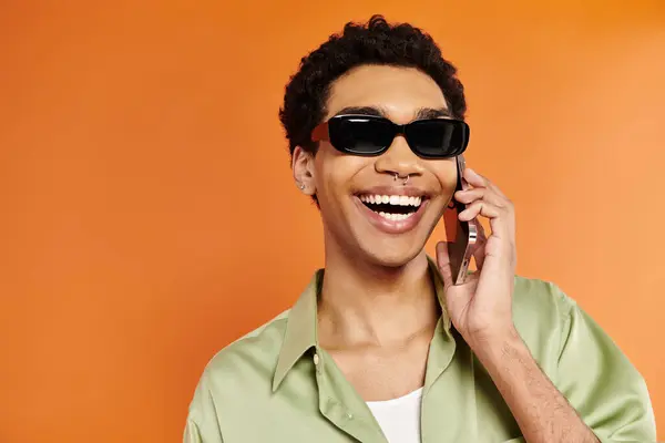 Alluring happy african american man with stylish sunglasses talking by phone on orange backdrop — Stock Photo