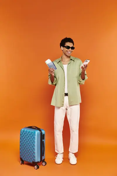 Alegre hombre afroamericano con gafas de sol con billete y pasaporte al lado de su maleta azul - foto de stock