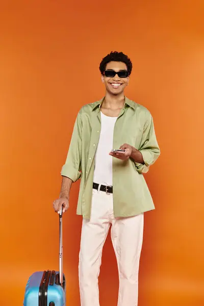 Alegre hombre afroamericano en traje casual con gafas de sol sosteniendo el teléfono junto a su maleta azul - foto de stock