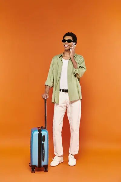 Alegre afroamericano hombre con gafas de sol de moda hablando por teléfono al lado es azul maleta - foto de stock