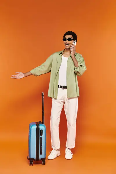 Joyeux homme afro-américain avec des lunettes de soleil à la mode parler par téléphone à côté est valise bleue — Photo de stock