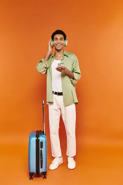 Alegre afroamericano hombre con auriculares sosteniendo el teléfono y mirando a la cámara cerca de la maleta - foto de stock
