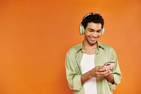 Joven feliz afroamericano hombre con auriculares usando su teléfono móvil en fondo naranja — Stock Photo