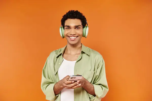 Alegre hombre afroamericano de moda con auriculares sosteniendo su teléfono inteligente y sonriendo a la cámara - foto de stock