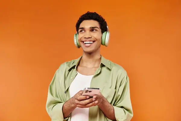 Elegante hombre americano africano alegre con auriculares sosteniendo el teléfono y mirando hacia otro lado, fondo naranja — Stock Photo