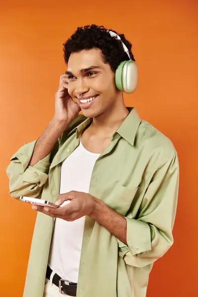 Hombre afroamericano alegre de moda con auriculares sosteniendo el teléfono y mirando hacia otro lado, fondo naranja — Stock Photo