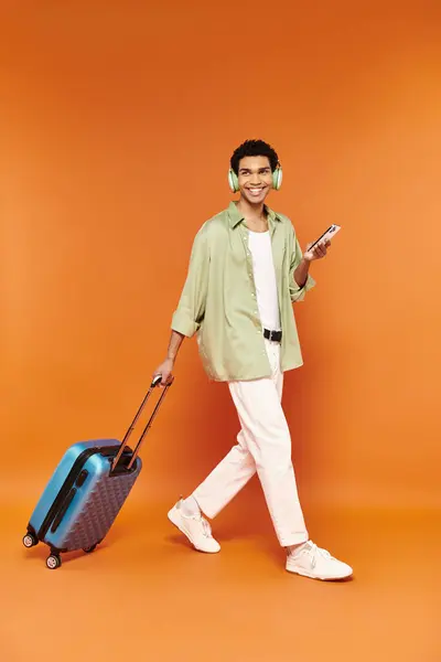 Gioioso uomo afroamericano con le cuffie che tiene il telefono e cammina con la sua valigia blu — Foto stock