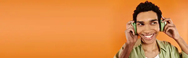 Allegro uomo afroamericano in abbigliamento accogliente con cuffie guardando altrove, sfondo arancione, banner — Foto stock