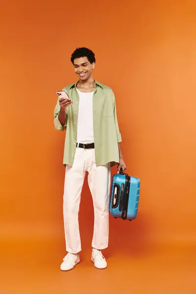 Gioioso giovane afroamericano guardando il suo cellulare e tenendo in mano la valigia, sfondo arancione — Foto stock